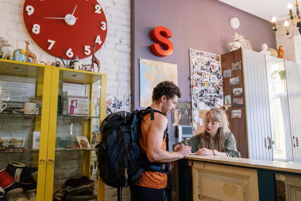 A backpacker at a hostel front desk getting assistance from a receptionist.