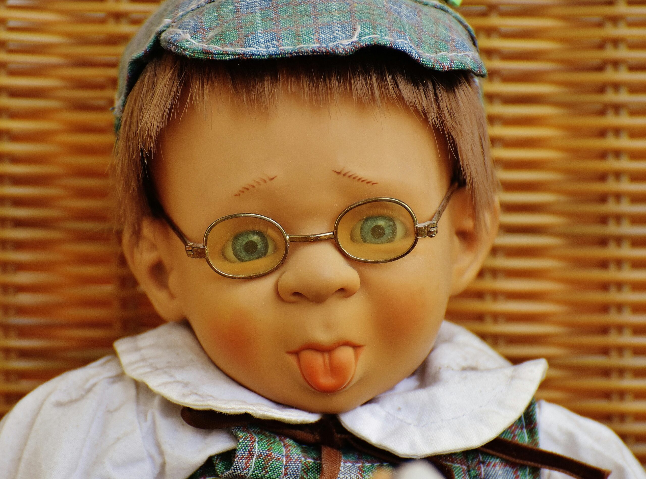 Adorable child-like doll wearing glasses and hat, playfully sticking out its tongue in a humorous portrait.