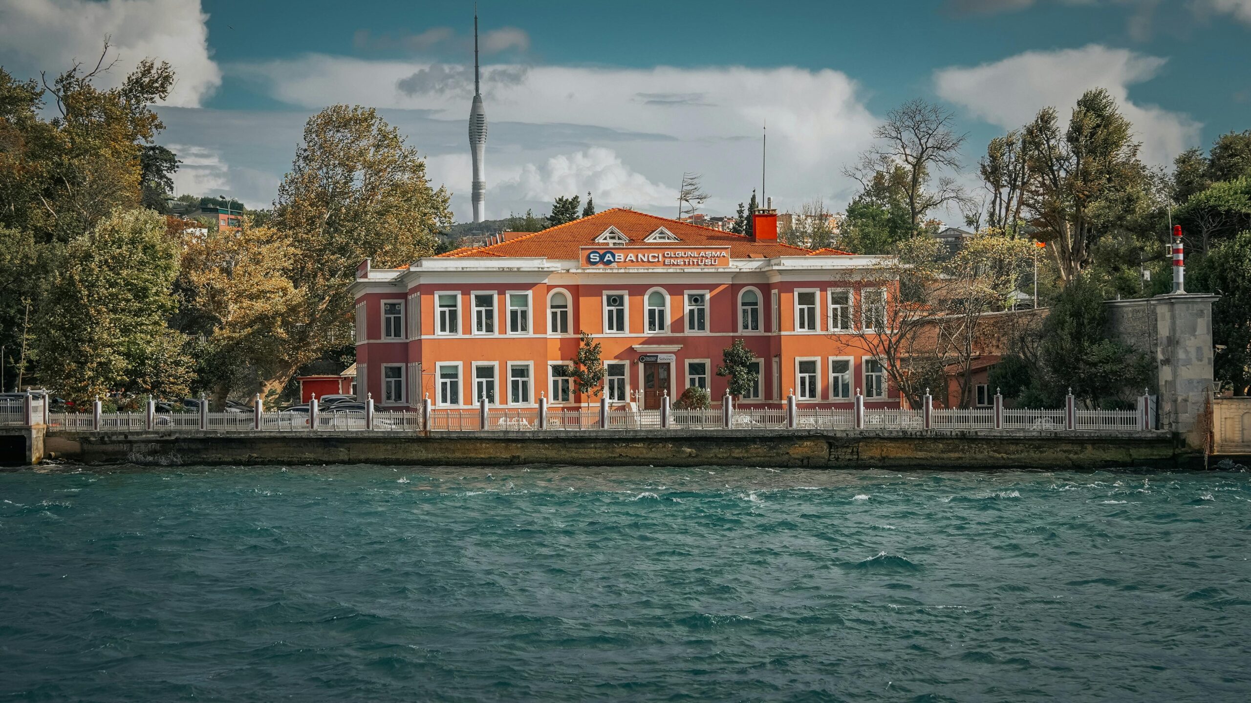 A charming historic building beside a body of water, framed by trees, with a prominent tower in the background.