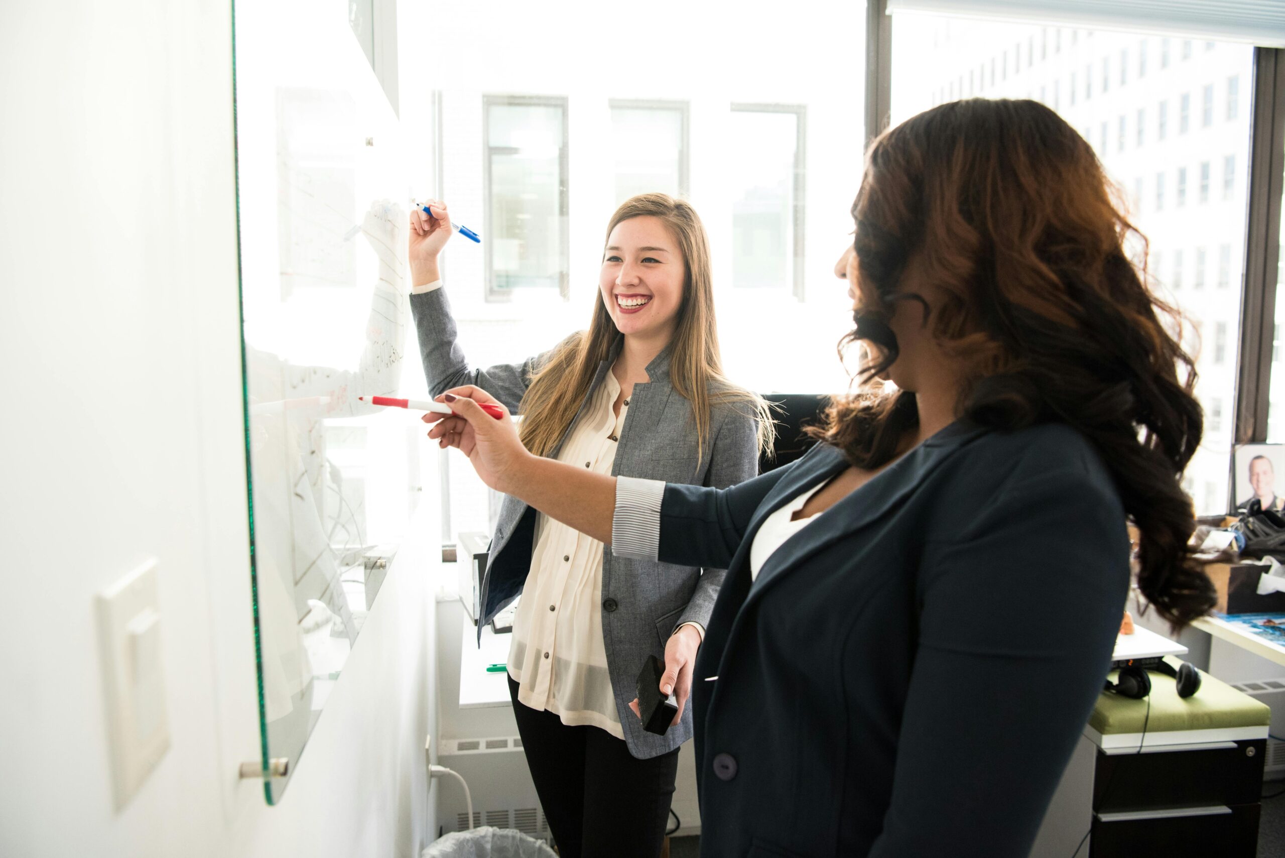 Two professional women discussing ideas on a whiteboard in a modern office setting.