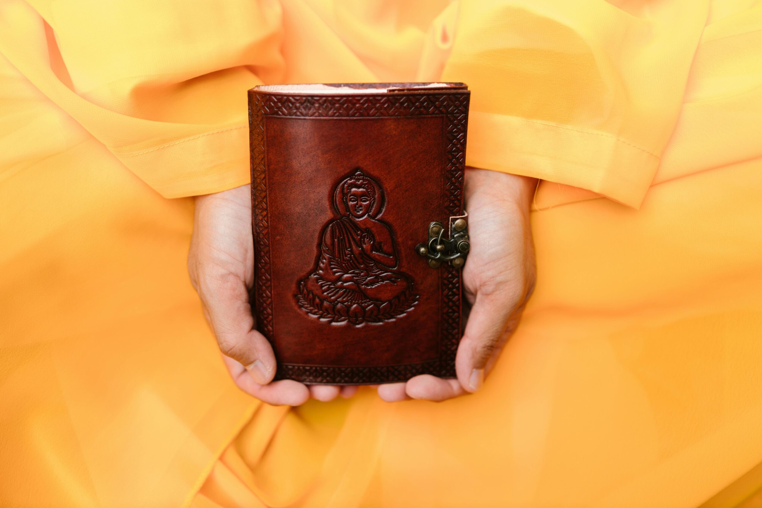 Close-up of hands in traditional attire holding a leather book with Buddha engraving.