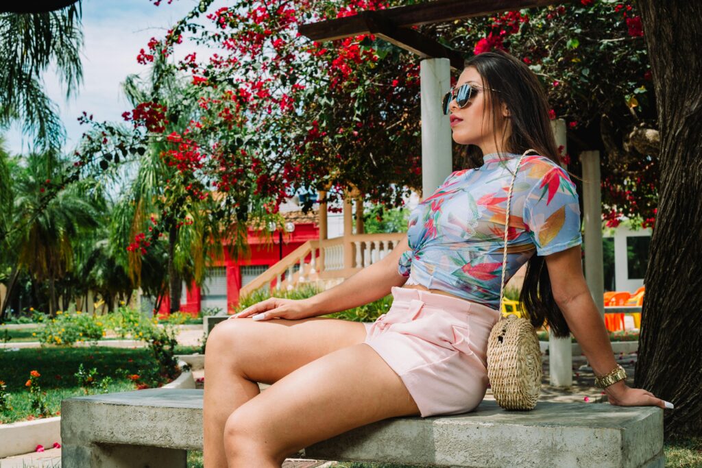 Fashionable woman in sunglasses sitting on a bench in a vibrant garden during summer.