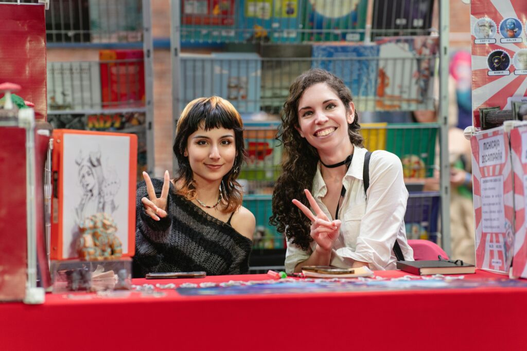 Two women at an anime convention in San José, Costa Rica, posing with peace signs, surrounded by art and comics.