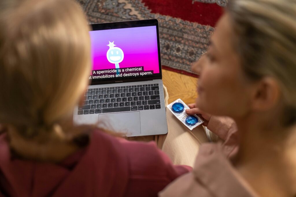 Two women watching an educational video on contraception and holding condoms indoors.