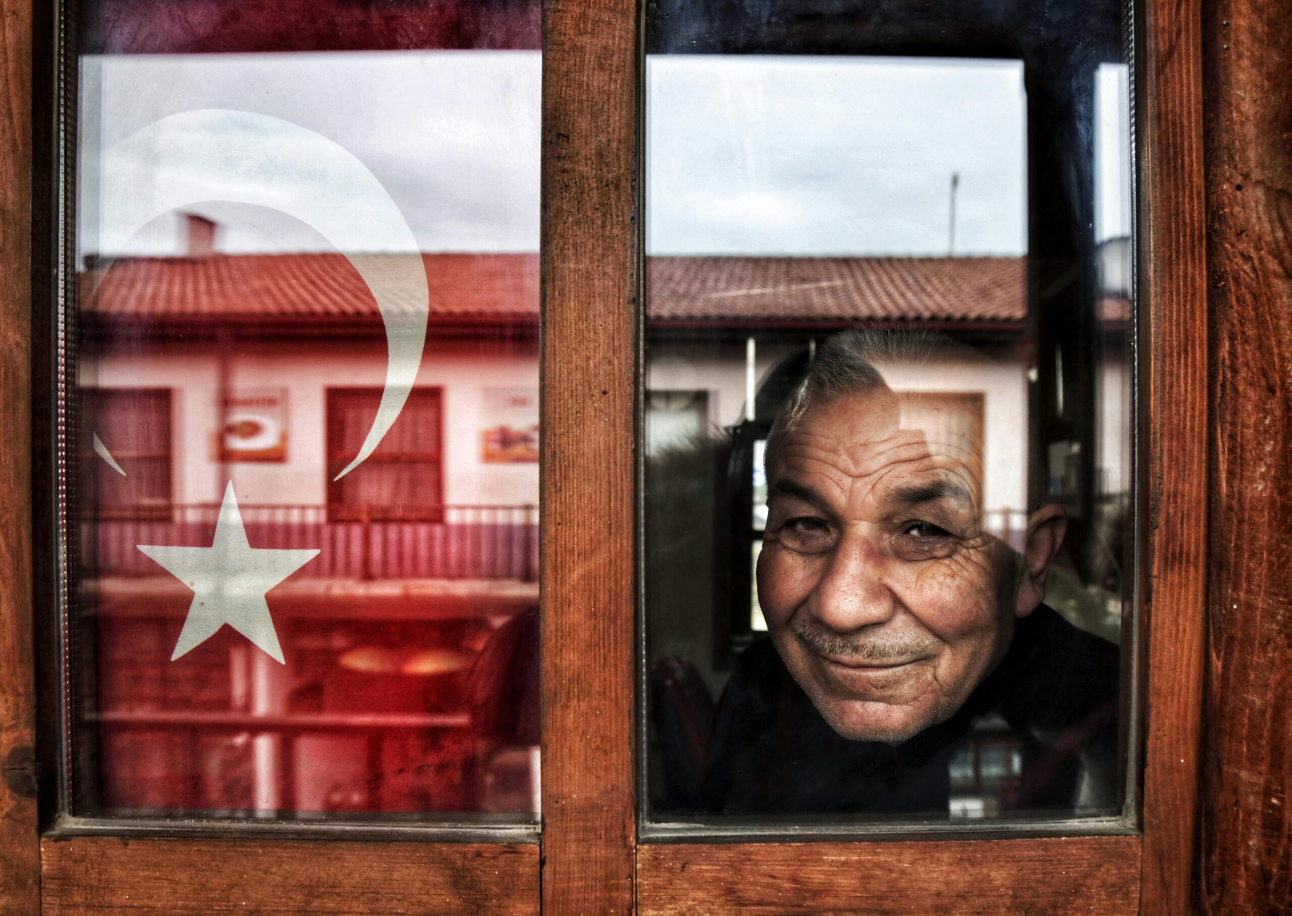 Portrait of an elderly man looking through a window with Turkish flag, Nallıhan, Türkiye.