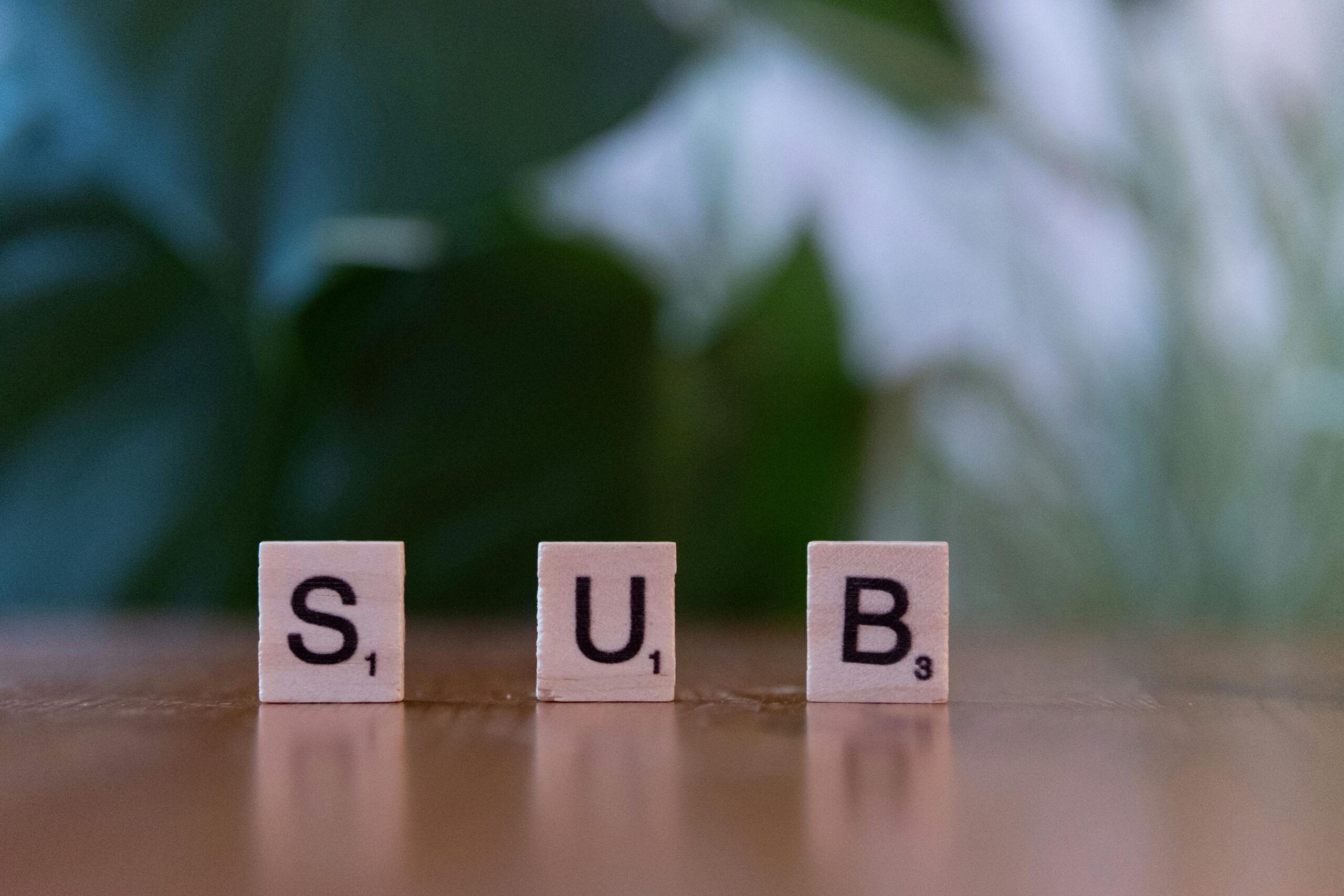 Three Scrabble tiles spelling 'SUB' on a wooden surface with blurred green background.