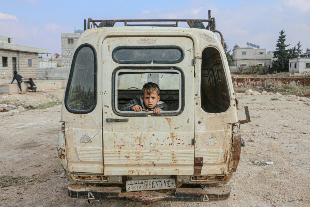 A young boy peers from an abandoned van in the desolate streets of Idlib, Syria, amidst war devastation.