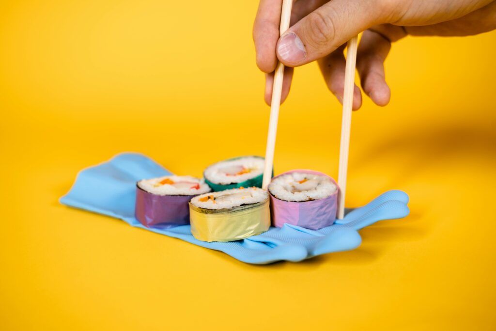 Sushi rolls wrapped in colorful foil with chopsticks on a yellow backdrop, highlighting pollution.