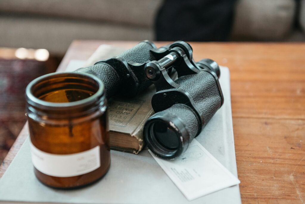 A close-up of vintage binoculars resting on a wooden table with a book and amber glass jar.