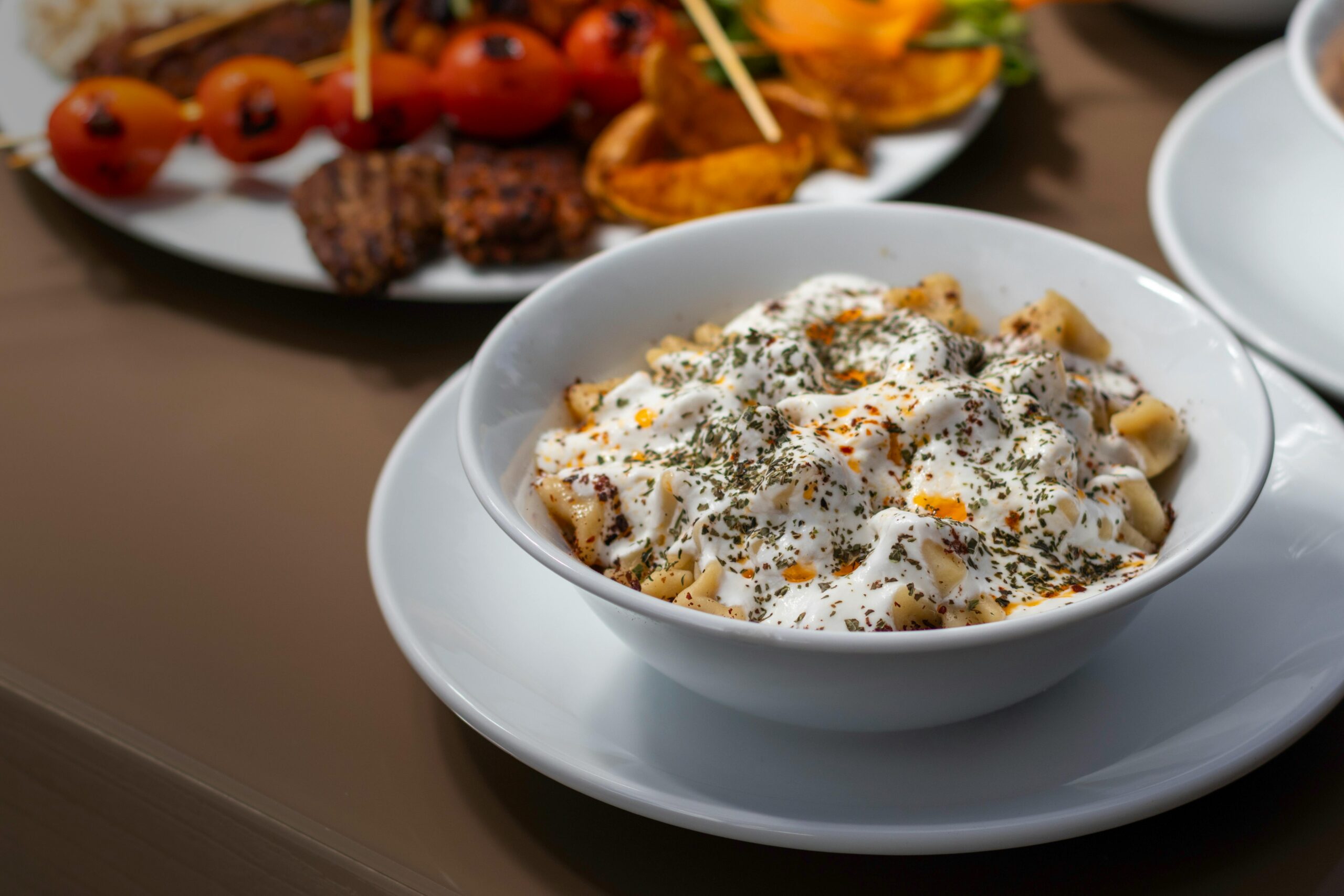 Turkish manti topped with yogurt and spices served with grilled vegetables on a sunny outdoor table.