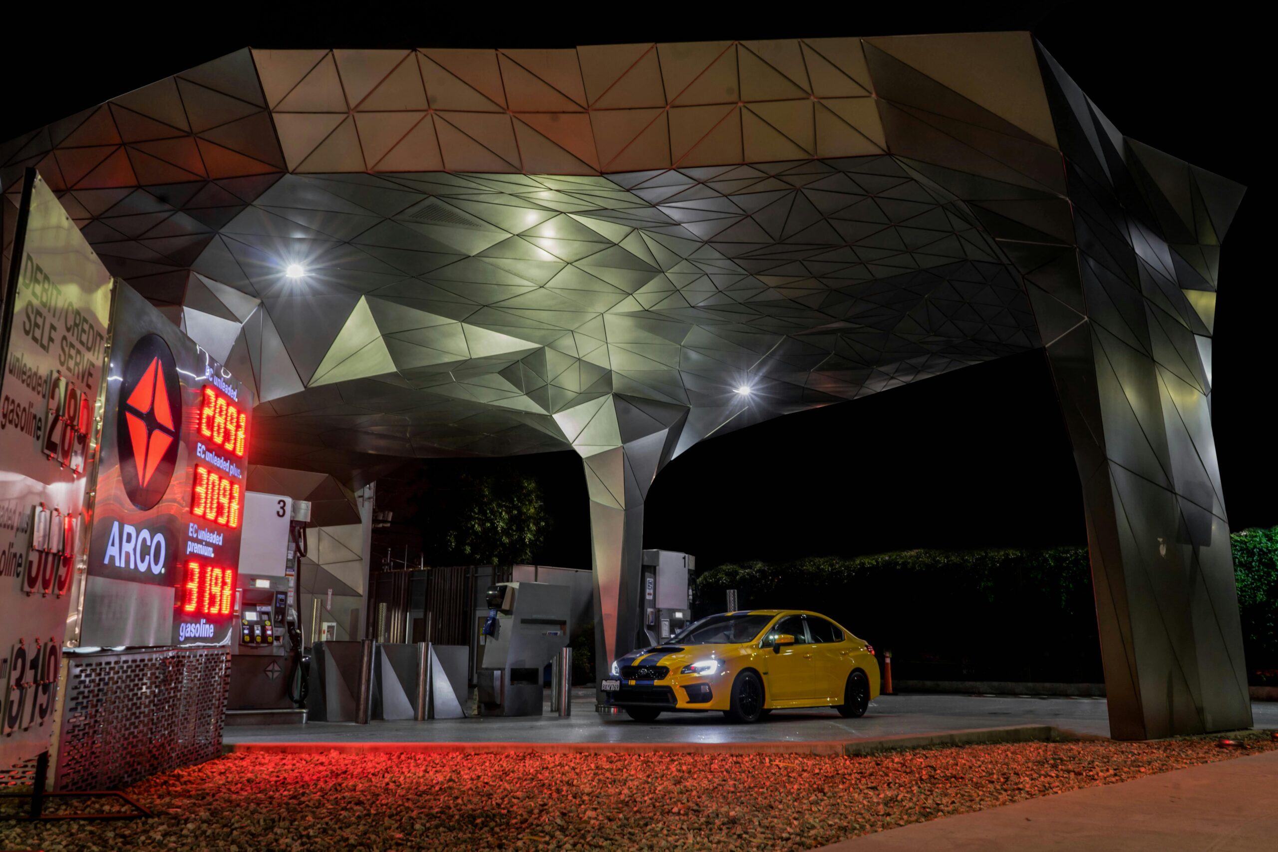 A futuristic gas station at night with illuminated canopy and vibrant lights, featuring a yellow car.