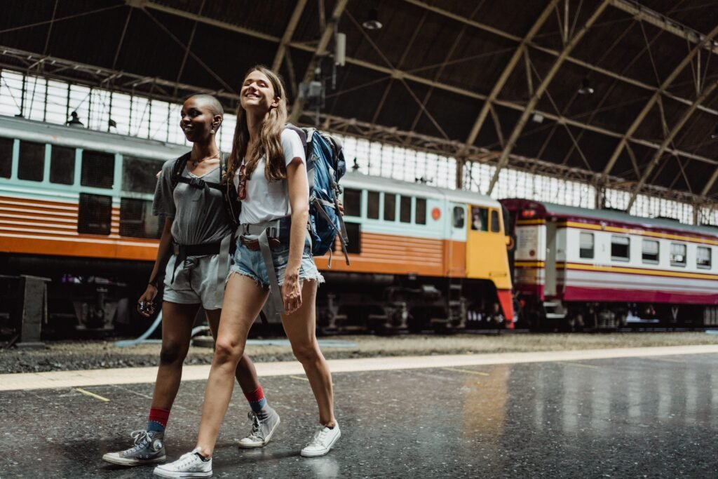 pexels-photo-4901993-4901993 Two women with backpacks walking and smiling at a railway station.